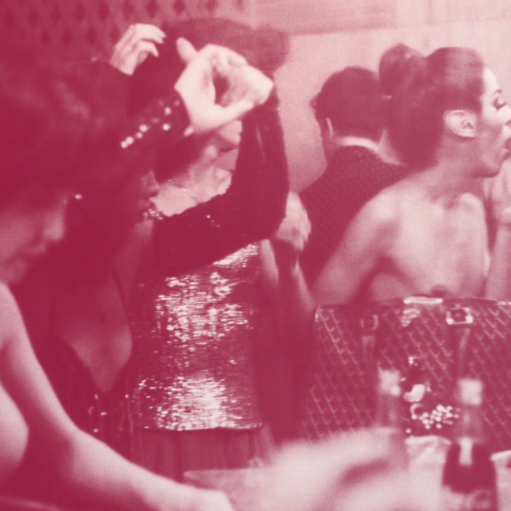 Fotografia preto e branco com filtro rosa de mulheres se maquiando em um camarim. Três delas estão com o braço em frente ao rosto pois arrumam os cabelos, enquanto uma mulher, que é uma figura mais central na fotografia, passa batom. Todas olham para o espelho.