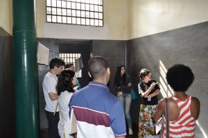 Foto de um grupo de pessoas dentro de uma cela olhando para uma educadora que aponta para uma parede cinza cheia de rasuras.