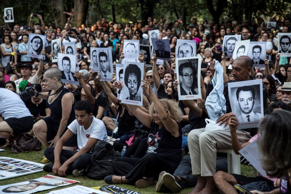 Foto colorida de pessoas reunidas em uma manifestação pacífica. Algumas delas seguram em frente ao corpo fotografias em preto e branco de desaparecidos e mortos políticos