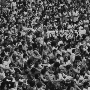 Foto em preto e branco de centenas de pessoas sentadas, bem próximas umas das outras. A imagem remete à uma manifestação. Ao entro, é possível ler em uma faixa a frase "Liberdades Democratas".