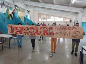 Foto colorida de alunos no pátio de uma escola. Quatro alunos seguram uma faixa feita com papel craft onde lemos " Ditadura Nunca Mais".