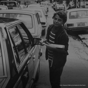 Foto em preto e branco de uma criança na rua, ao lado de um carro antigo. Ela segura na mão uma caixa de chiqueletes, veste um moletom, uma blusa listrada e calça jeans.