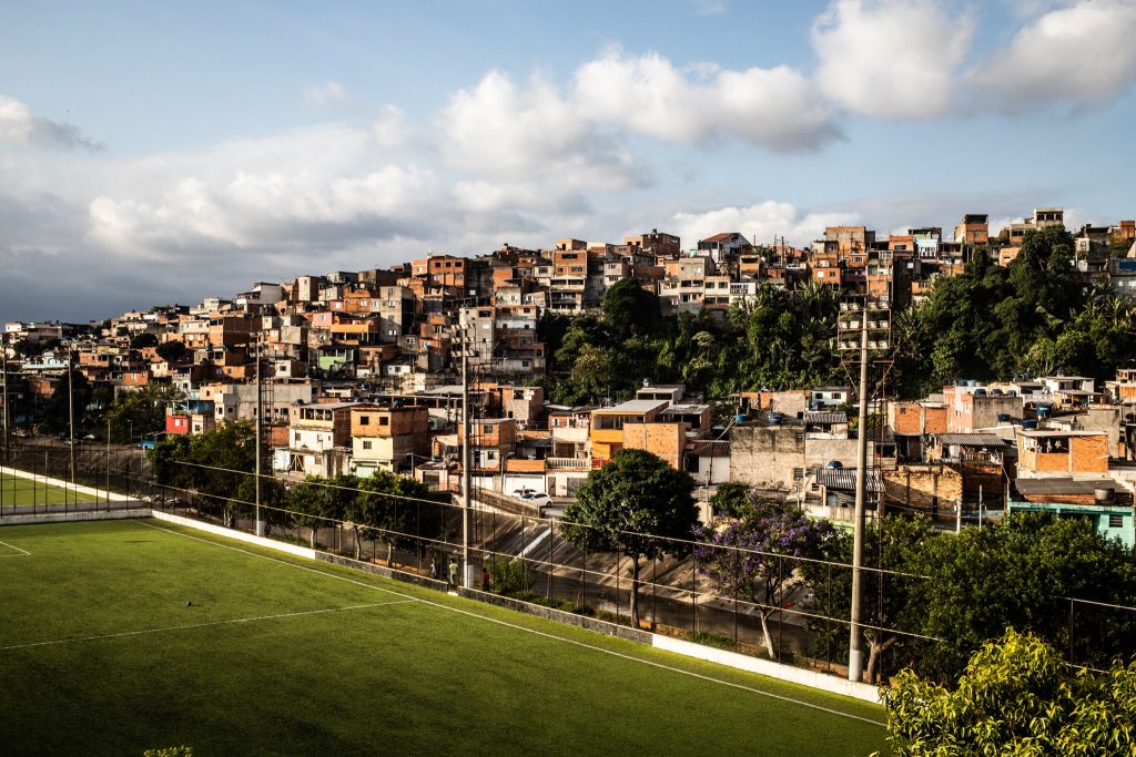 Foto colorida de paisagem. Em primeiro plano, um campo de futebol gramado. Ao fundo, ocupando a montanha, casas com tijolos aparentes e um céu azul com nuvens ao fundo.