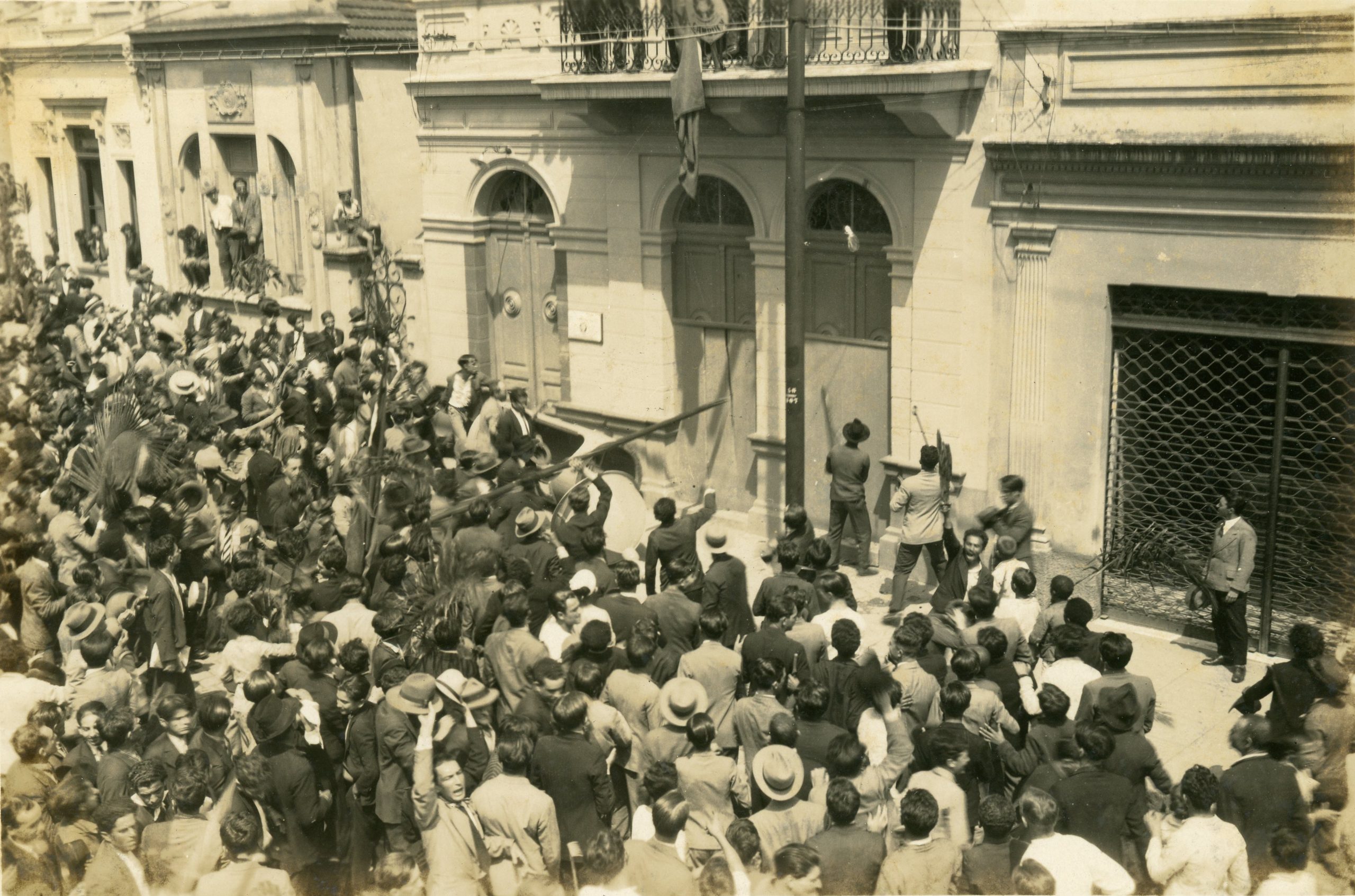 Foto em preto e branco de uma grande multidão de homens tentando invadir um prédio. Na parte central da imagem vemos que parte deste grupo segura uma barra de ferro que está sendo pressionada sobre a porta de um edifício.