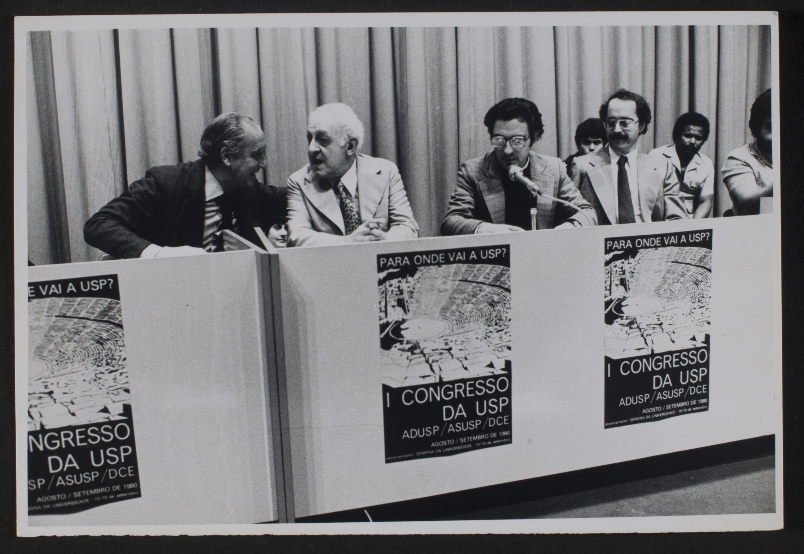 Foto preto e branco com a mesa de debates e 4 homens maduros de terno e gravata sentados. Em frente a mesa figura 3 cartazes iguais onde se lê: I Congresso da USP. Ao fundo da mesa 3 homens mais jovens sentados de camisa.