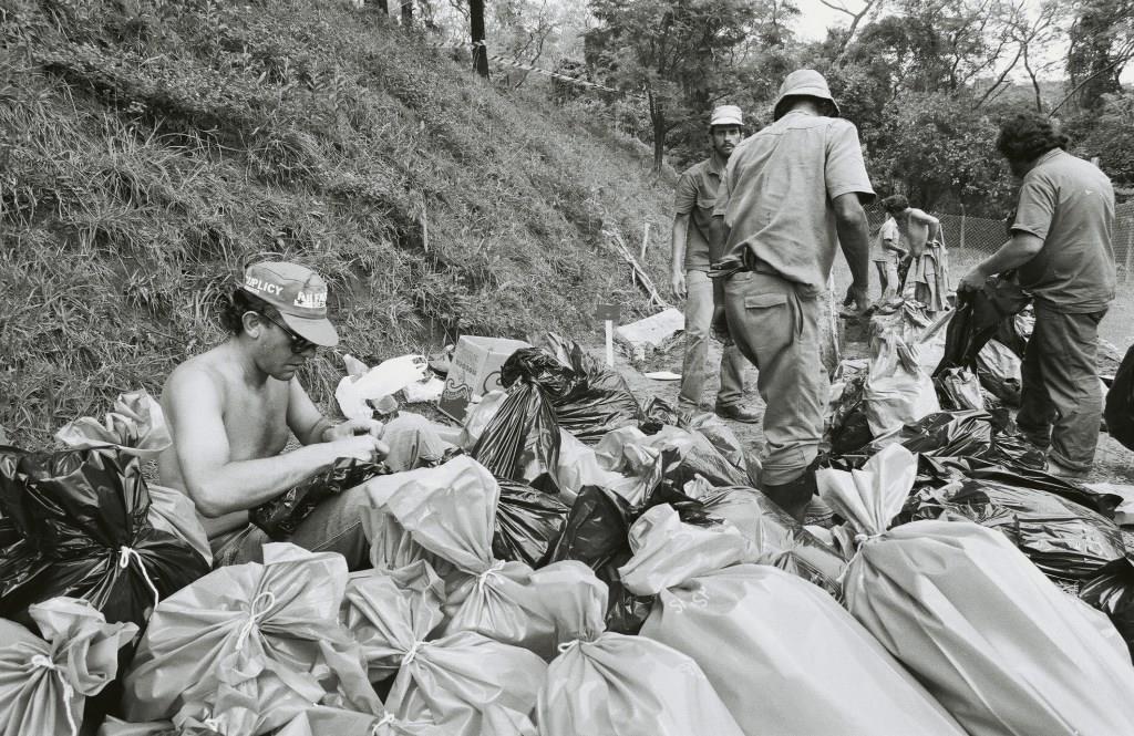 Foto em preto e branco de vários sacos empinhados. Por entre eles um homem sem camisa mexe em um enquanto dois soldados transitam entre eles. Um quarto homem empilha mais um saco no local.