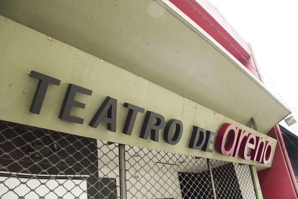Foto colorida de detalhe de letreiro em fachada. Lemos "Teatro de" com letras pretas e "arena" com letras vermelhas.