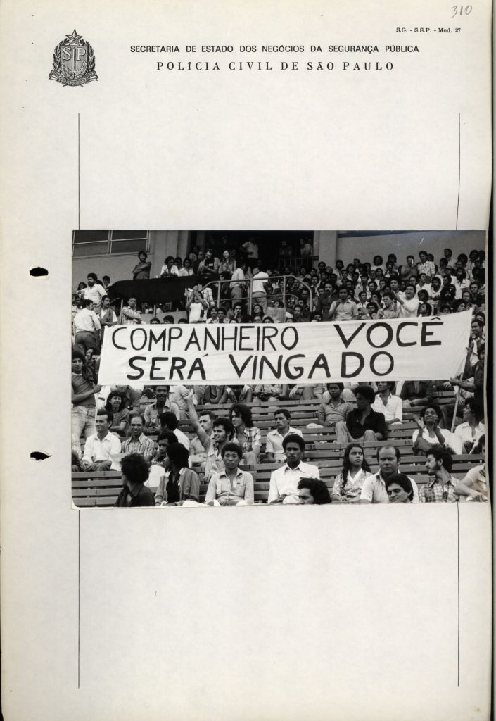 Foto em preto e branco de várias pessoas sentadas em arquibancada de estádio de futebol. Elas seguram uma faixa onde lê-se “Companheiro você será vingado”.