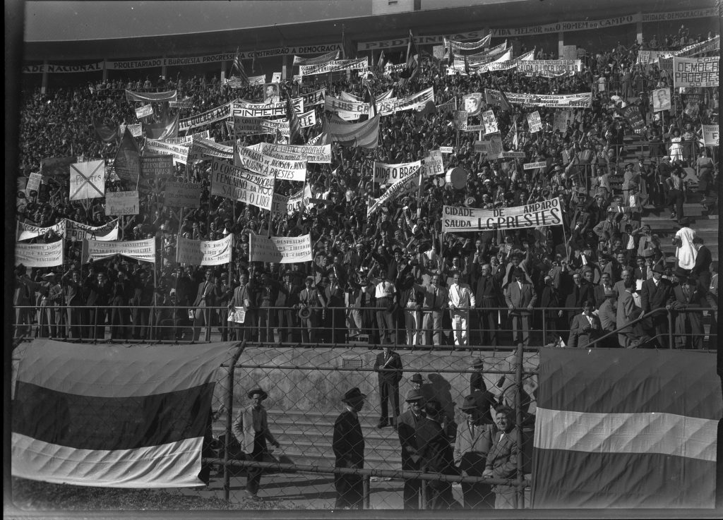 Foto em preto e branco de uma arquibancada de estádio de futebol lotada. A maioria das pessoas seguram faixas.