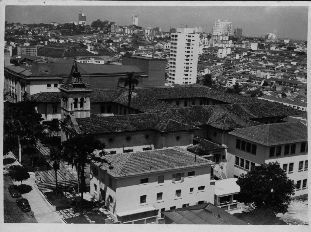 Foto em preto e branco de paróquia com torre surgindo de um edifício de 3 andares à esquerda. No centro estes edifícios de 3 andares formando um quadrado e no fundo várias casas e prédios residenciais.
