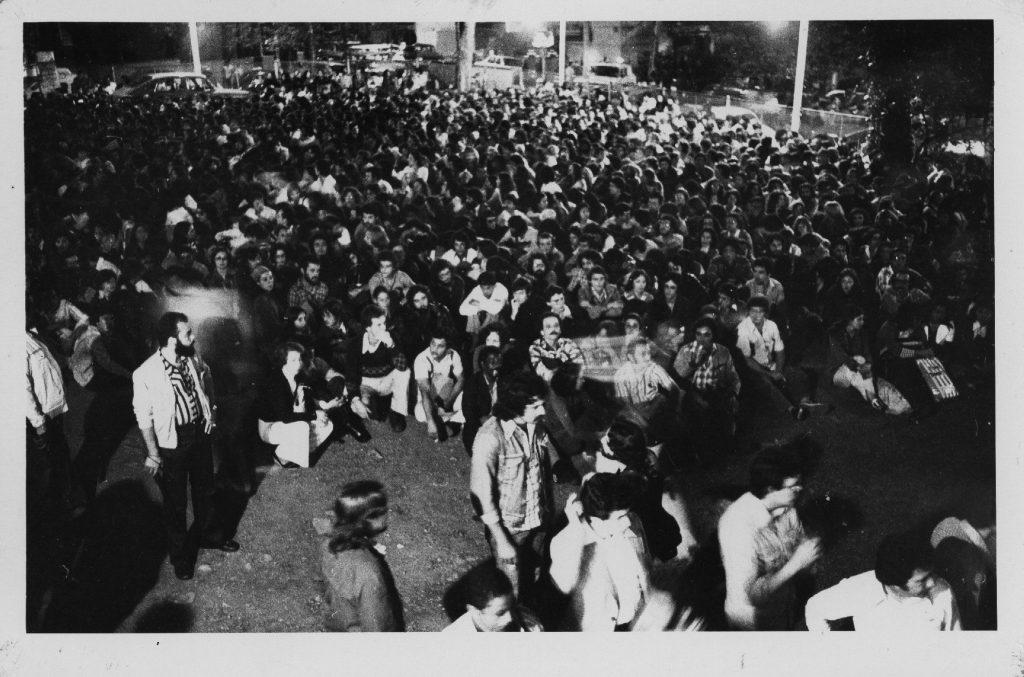 Foto em preto e branco de várias pessoas sentadas no chão de um vão e algumas pessoas em pé no primeiro plano da imagem
