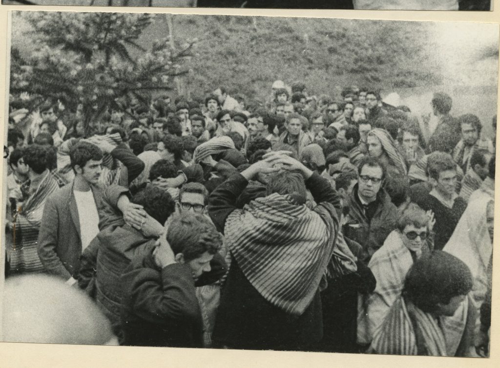 Foto em preto e branco de jovens aglomerados, a maioria deles com as duas mãos sobre a nuca em sinal de rendição.