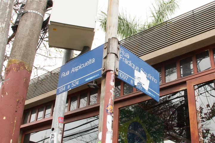 Foto colorida com detalhe da placa azul de rua que coloca-se em uma esquina. Nela podemos ler em branco os nomes: Rua Aspicuelta, na placa da esquerda, e rua Fradique Coutinho, na placa da direita. Ao fundo uma porta de vidro grande.