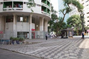 Foto colorida de espaço térreo de prédio branco,circular onde podemos ver 4 colunas retangulares. A frente do prédio um calçadão com desenhos da forma que possui o Estado de São Paulo formando um mosaico preto e branco.