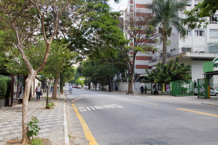 Foto colorida de uma avenida larga e arborizada com prédios em seu entorno. A avenida está sem carros e com poucas pessoas na calçada. Na lateral esquerda é possível ver um ponto de ônibus atual, de vidro e ferro.