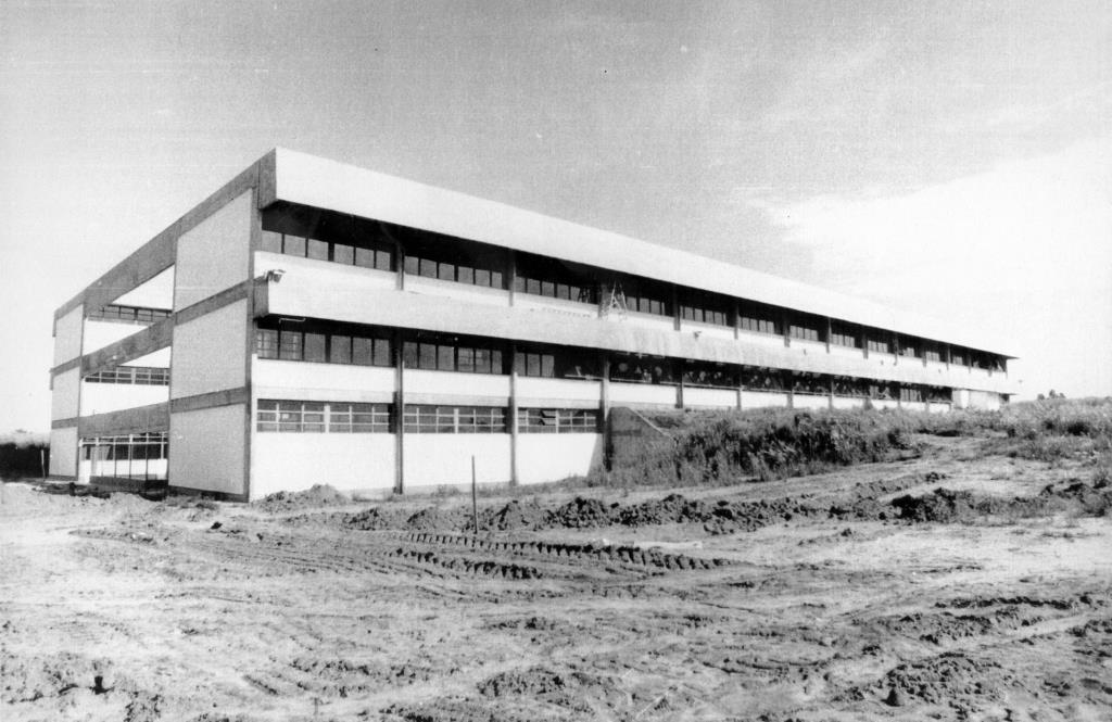 Foto em preto e branco de um grande prédio de 3 pavimentos em construção. A fachada é branca com janelas seguidas. À frente dele há o terreno mexido de terra com marcas de trator