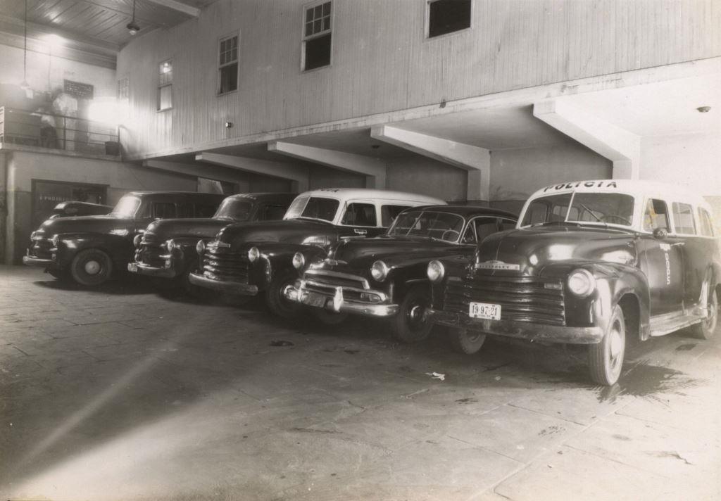 Fotografia em preto e branco de um estacionamento coberto com 5 carros dos anos 50. Todos são escuros sendo que 2 deles (o do canto direito e o do centro) são viaturas, pois tem a cobertura branca e tem o escrito “polícia” na junção entre o para-brisa e a cobertura.