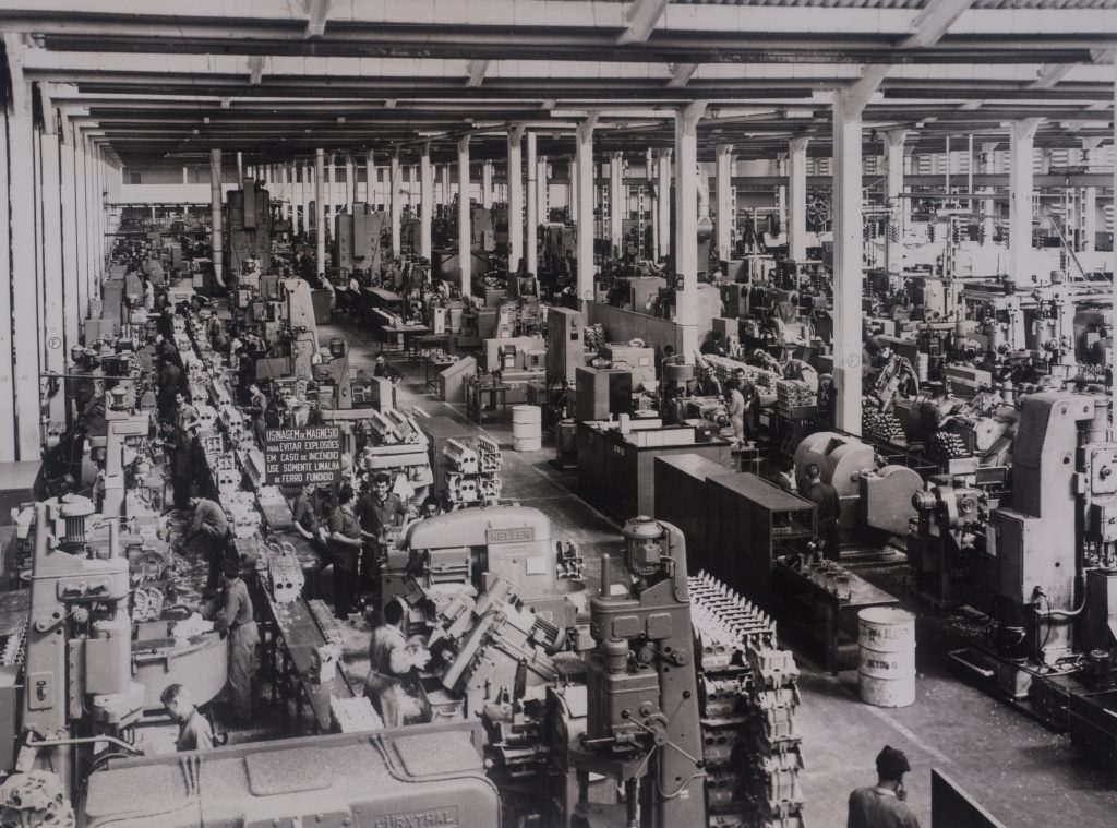 Foto em preto e branco de um enorme galpão repleto de máquinas. Na imagem as pessoas aparecem pequenas e se mesclando por entre os equipamentos. No canto esquerdo vemos uma esteira que carrega produtos.