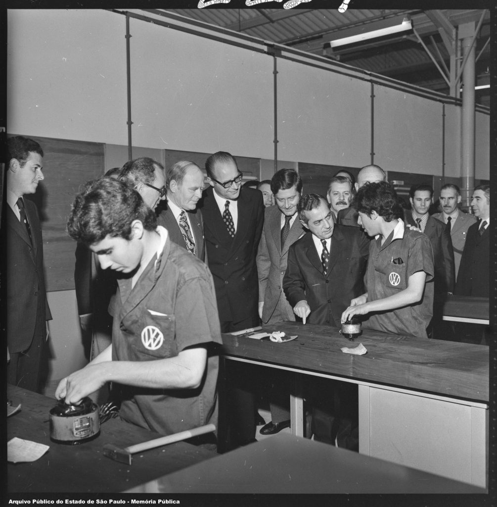 Foto em preto e branco de 2 homens uniformizados com camisa com logotipo da Volkswagen no bolso trabalhando em suas mesas. Na mesa central, vários homens de terno e gravata conversam com o homem que trabalha.