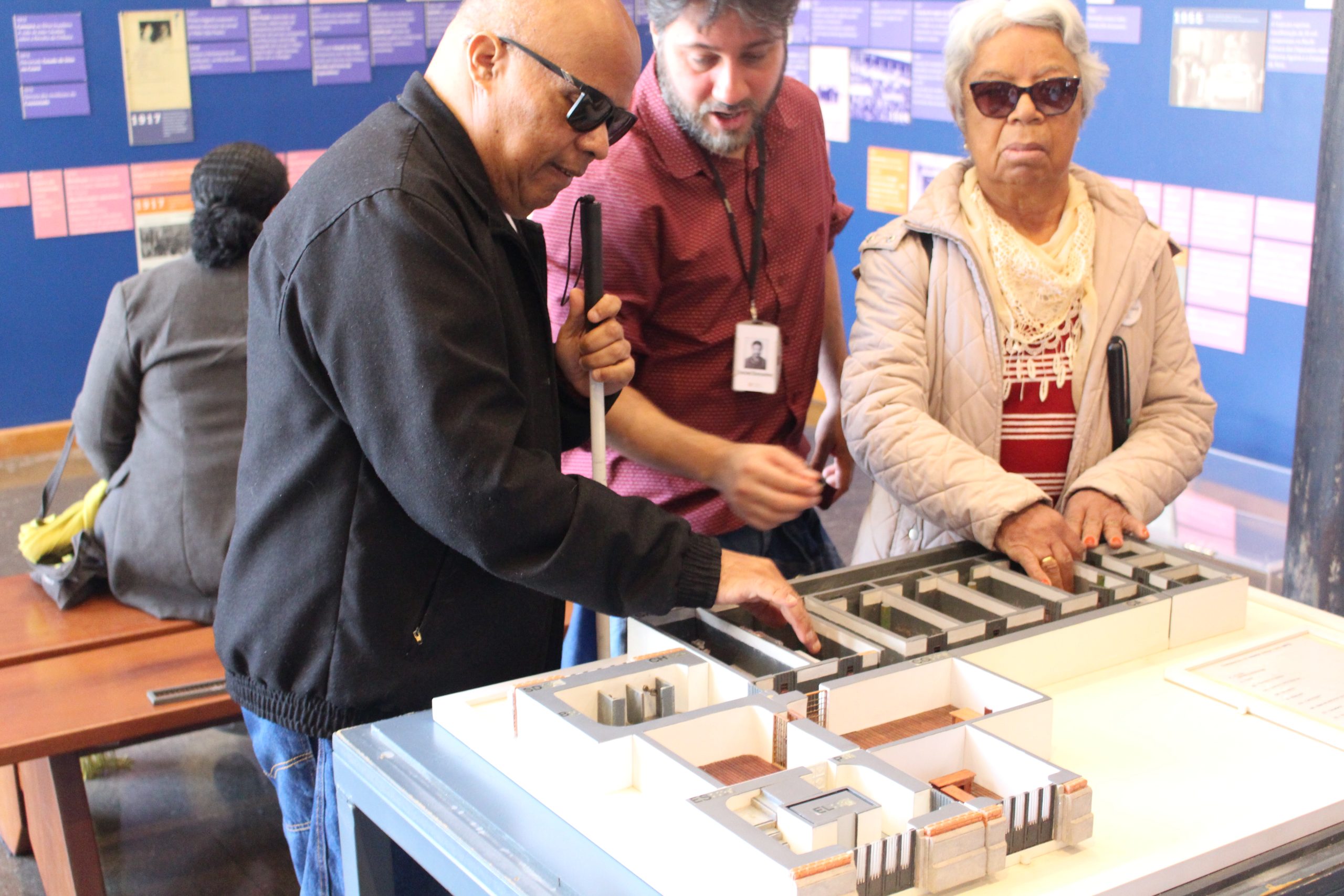 Imagem mostra uma visita mediada por um educador e um intérprete do Programa Memorial Para Todos, no Memorial da Resistência de São Paulo, com visitantes com deficiência visual. A maquete em alto relevo, que integra a exposição permanente, está sendo apresentada. Ela permite que o público tenha uma noção do que restou do espaço carcerário que abrigou, por mais de quatro décadas, o Departamento Estadual de Ordem Política e Social de São Paulo (Deops/SP).