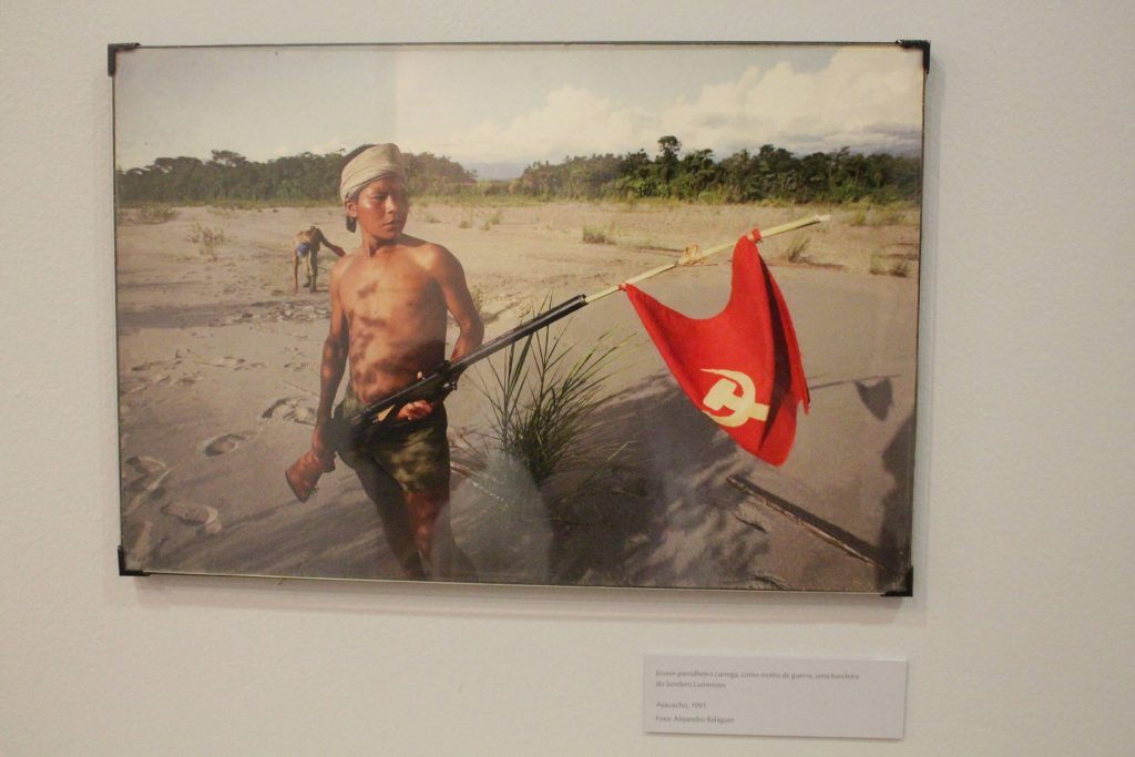 Foto de um quadro fotográfico colorido com a figura de um homem sem camisa e usando bermuda, com um lenço na cabeça e segurando uma bandeira vermelha com o desenho de uma foice e um martelo em branco.