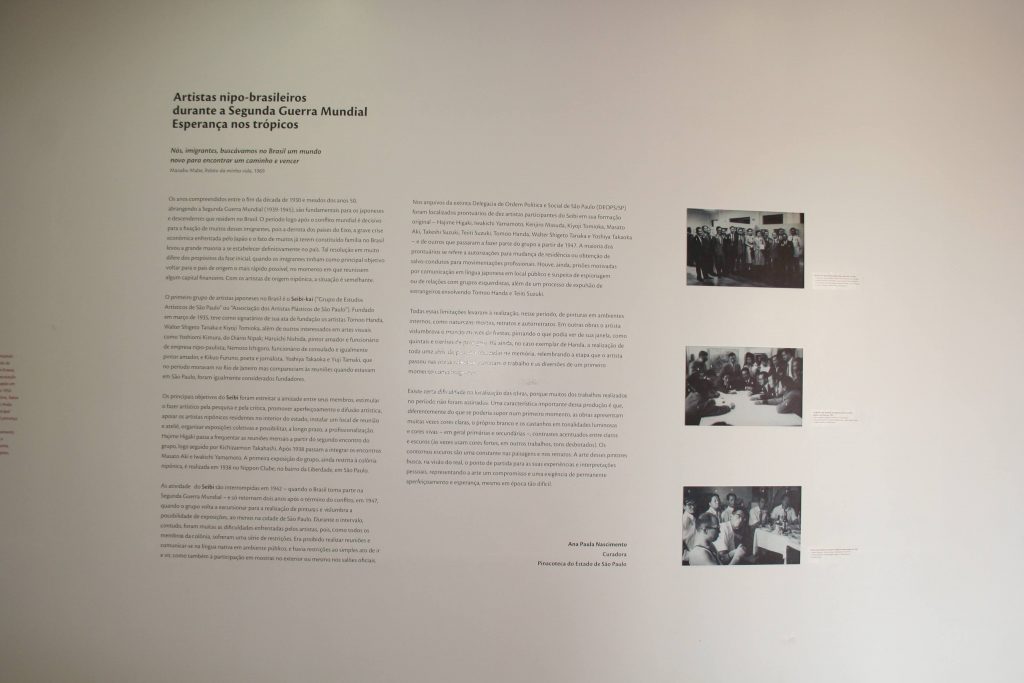 Foto de parede branca com 3 fotos em preto e branco plotadas de pessoas reunidas, no canto direito. Ocupando o centro e o canto esquerdo vemos um texto cujo o título diz: “ Artistas nipo-brasileiros durante a Segunda Guerra Mundial: Esperança nos trópicos”.