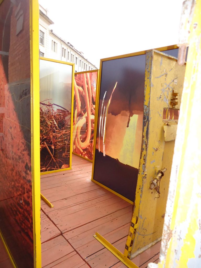 Foto de painéis de metal pintado de amarelo com fotos coloridas de espaços em ruínas sobre um tablado de madeira.
