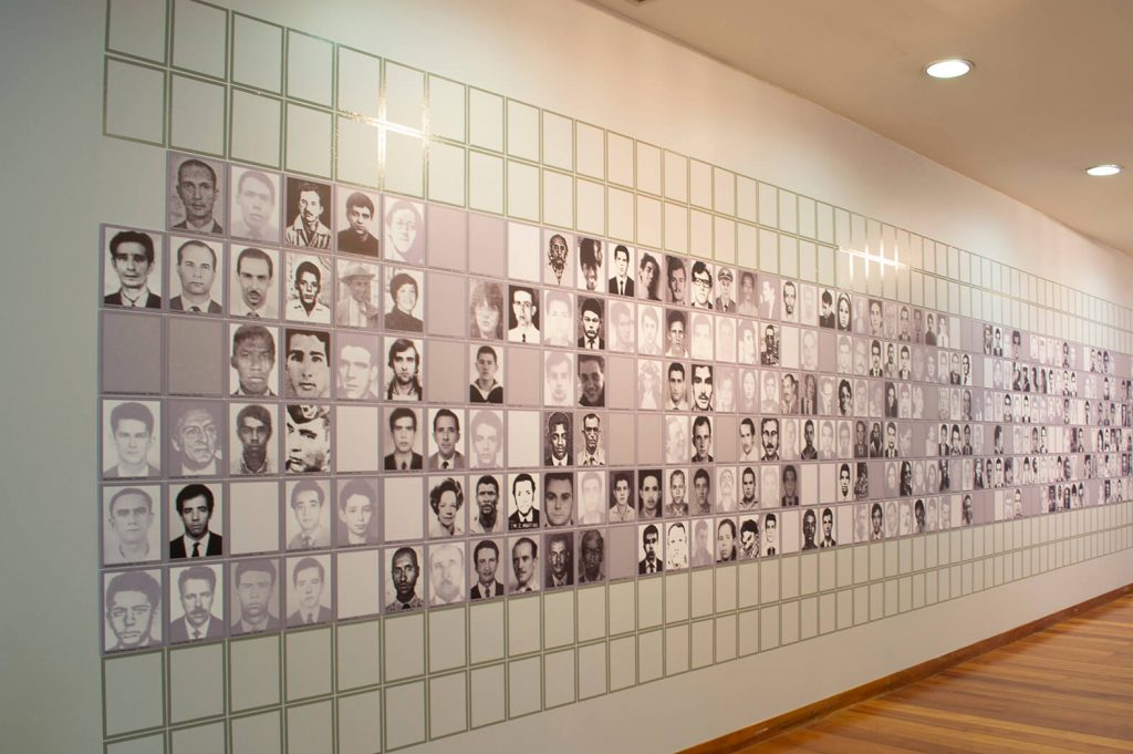 Foto de parede branca em que parece plotado muitas fotos 3x4 de homens e mulheres em preto e branco, formando um quadriculado. Neste quadriculado, por entre as fotos, aparecem alguns espaços vazios.