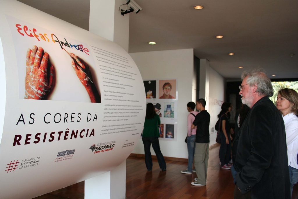 Foto colorida de painel onde lemos “Elifas Andreato - As Cores da resistência''. Um homem e uma mulher observam este painel, enquanto outras pessoas circulam pela exposição.