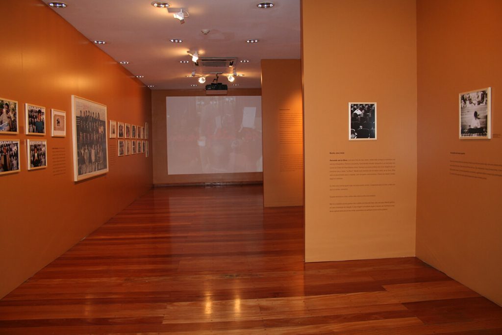 Foto colorida de sala expositiva com quadros com fotografias de pessoas pregados em paredes beges, piso de madeira e uma projeção com vídeo ao fundo.