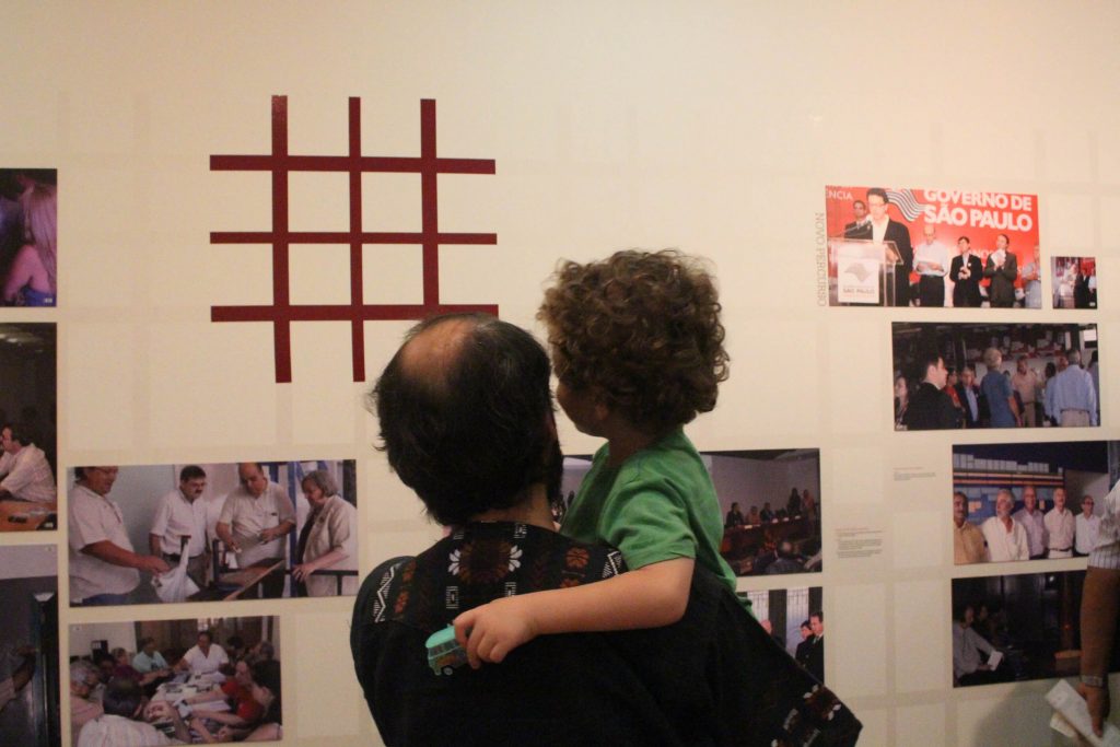 Fotografia colorida com um homem segurando uma criança no colo, ambos estão de costas para a foto e estão olhando para um parede branca com várias fotografias espalhadas ao redor da logomarca do Memorial da Resistência.