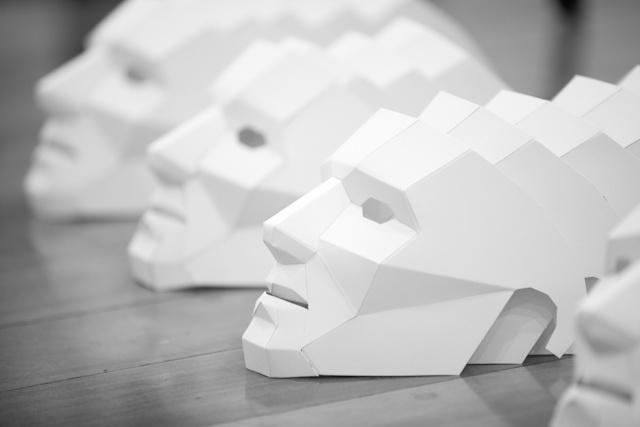 Fotografia em preto e branco de várias máscaras iguais feitas em papel branco enfileiradas uma à frente da outra.