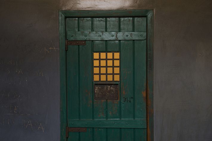 Foto horizontal de uma porta verde de uma das celas da Exposição de longa duração. As paredes são de cimento escuro com inscrições dos ex-presos.