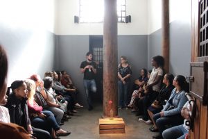 Foto de um grupo de mulheres sentadas nas laterais de uma cela, enquanto um educador fala. Elas olham para ele. No meio da foto vemos um caixote de madeira com um cravo vermelho dentro de uma embalagem de vinagre com água em frente a uma pilastra.