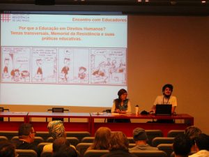 Fotografia de um homem e uma mulher em uma mesa com um notebook na frente do rapaz. Eles estão em um auditório com pessoas observando uma apresentação projetada na parede de uma tirinha da personagem Mafalda.