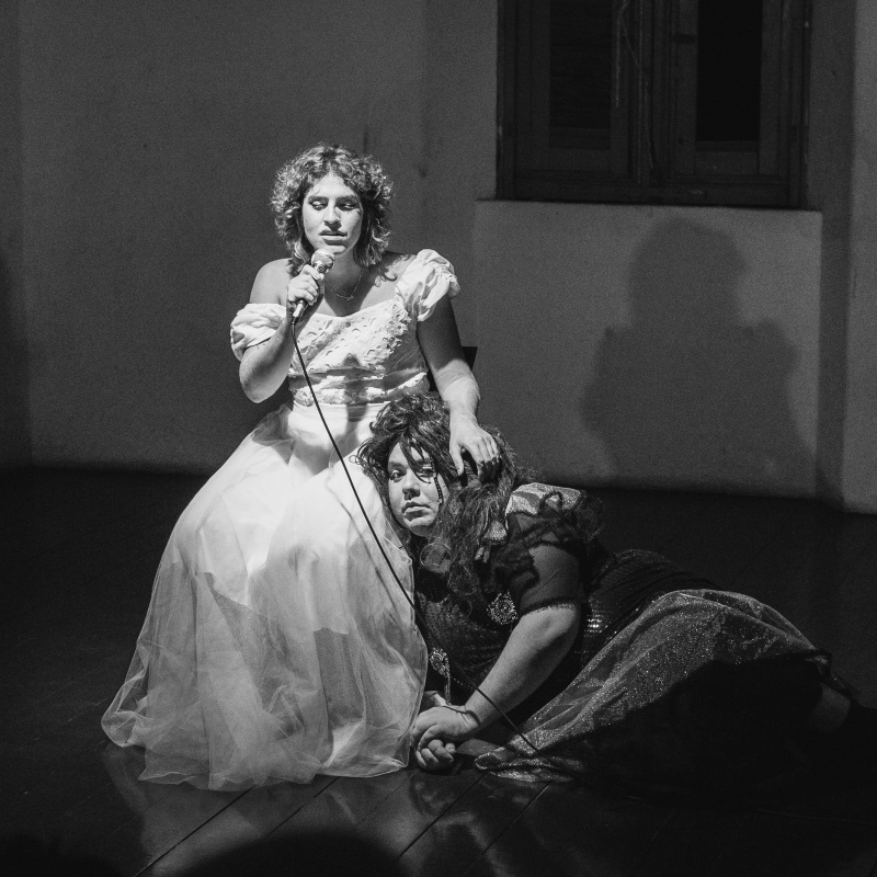 Foto em preto e branco de uma mulher sentada com um vestido branco segurando um microfone. Ao lado uma mulher de cabelos e vestes pretas encostada em sua perna, com uma expressão triste.