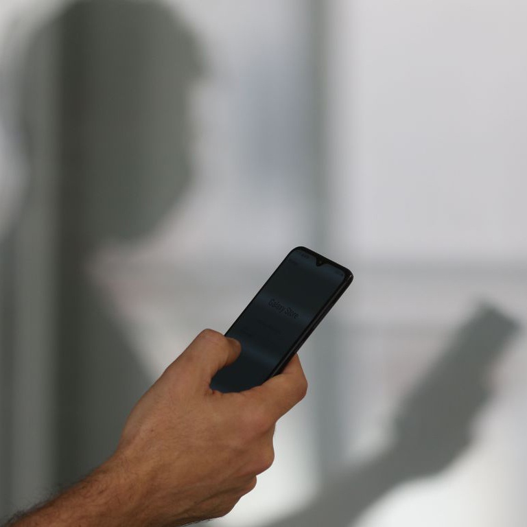 Fotografia colorida de um homem branco segurando um celular. Ao fundo vemos a sua sombra refletida na parede de cor cinza.