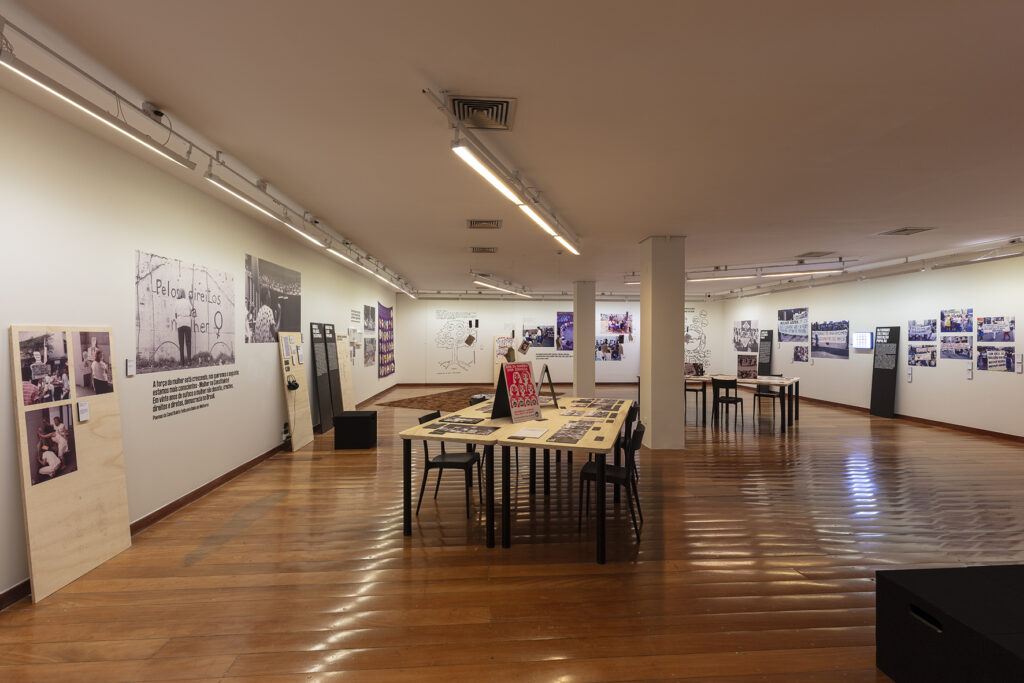 Foto colorida de espaço expositivo amplo com fotos coploridas plotadas nas paredes. No centro da imagem vemos uma mesa com fotos em seu tampo e cadeiras no seu entorno.