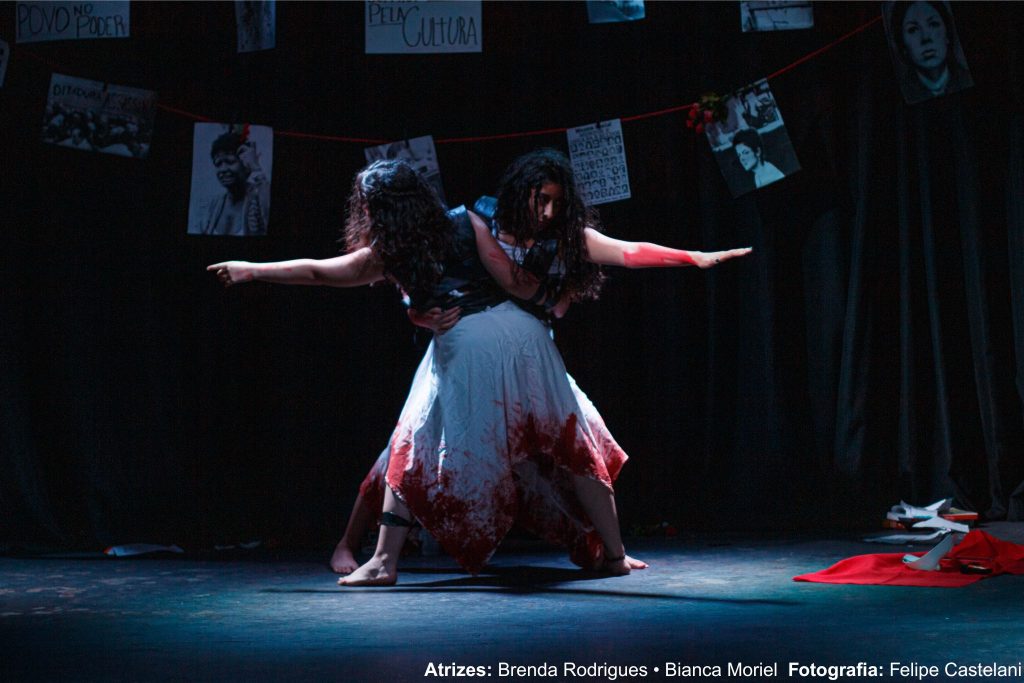 Foto colorida da peça de teatro "Inimigas Públicas", onde duas atrizes aparecem no centro do palco, uma de frente para a outra com o braço esquerdo erguido, de vestido branco com manchas vermelhas.