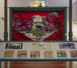 Foto colorida de sala de exposição com piso de madeira e colunas de ferro. No centro da sala vemos uma vitrine com páginas de jornal expostas e ao fundo uma bandeira do brasil estilizada em vermelho.