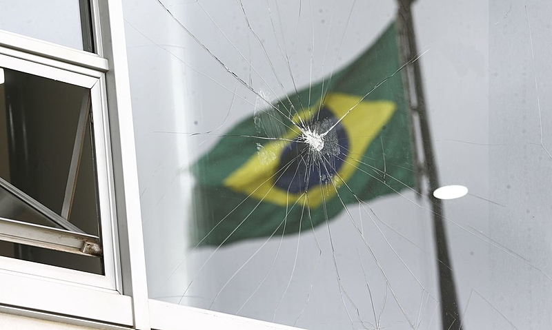 Foto colorida de uma janela com o vidro quebrado. Vê-se a bandeira do Brasil hasteada no reflexo da janela, de modo que as rachaduras no vidro estão no meio da bandeira.