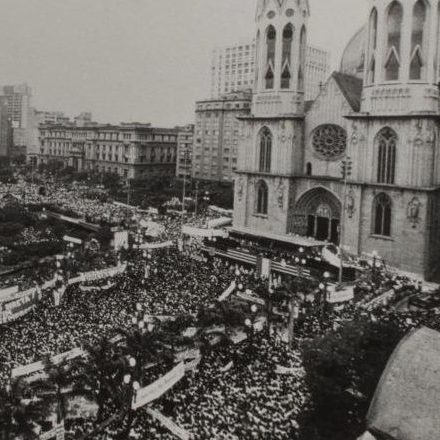 Fotografia em preto e branco de uma grande multidão aglomerada em frente a Catedral da Sé em São Paulo. A fotografia foi tirada do alto e podemos ver toda a área está repleta de pessoas, sendo que muitas delas seguram faixas. Ao fundo da imagem vemos prédios. A Igreja ocupa a lateral direita da foto e tem 2 torres de 92 metros de altura cada com janelas de arco ogival. Acima da porta de entrada, também em arco ogival, há uma rosácea (um vitral redondo). No local da escadaria em frente a igreja está montado um palanque feito em metal.