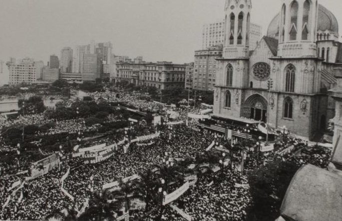 Fotografia em preto e branco de uma grande multidão aglomerada em frente a Catedral da Sé em São Paulo. A fotografia foi tirada do alto e podemos ver toda a área está repleta de pessoas, sendo que muitas delas seguram faixas. Ao fundo da imagem vemos prédios. A Igreja ocupa a lateral direita da foto e tem 2 torres de 92 metros de altura cada com janelas de arco ogival. Acima da porta de entrada, também em arco ogival, há uma rosácea (um vitral redondo). No local da escadaria em frente a igreja está montado um palanque feito em metal.