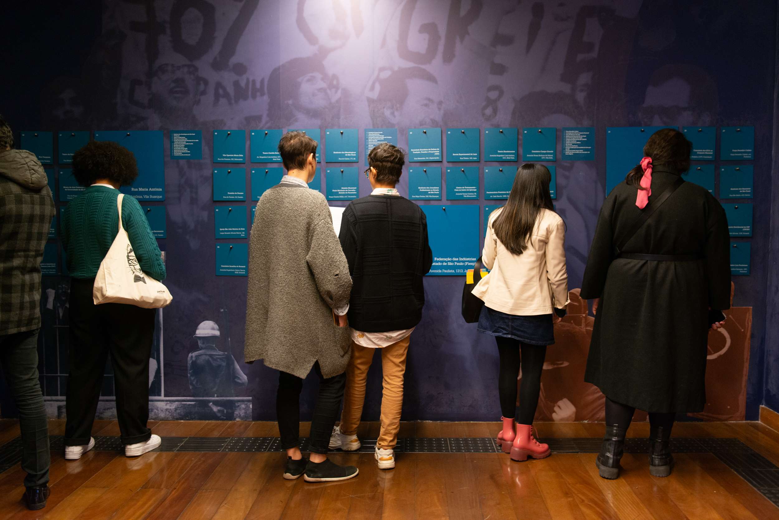 Pessoas de costas para câmera, observando pesquisa do Espaço de memórias que faz parte da exposição de Longa Duração do Memorial da Resistência de São Paulo que apresenta ao público o que restou do espaço carcerário do (DEOPS/SP) e a memória da resistência e violência causada pelo regime civil militar na ditadura.