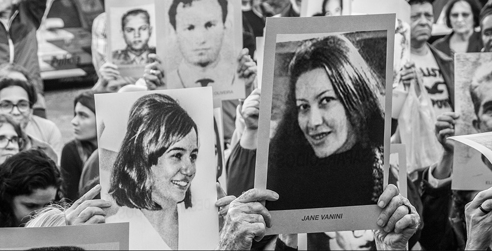 A imagem mostra uma manifestação com pessoas segurando cartazes em preto e branco com fotos e nomes de desaparecidos políticos da ditadura civil-militar brasileira