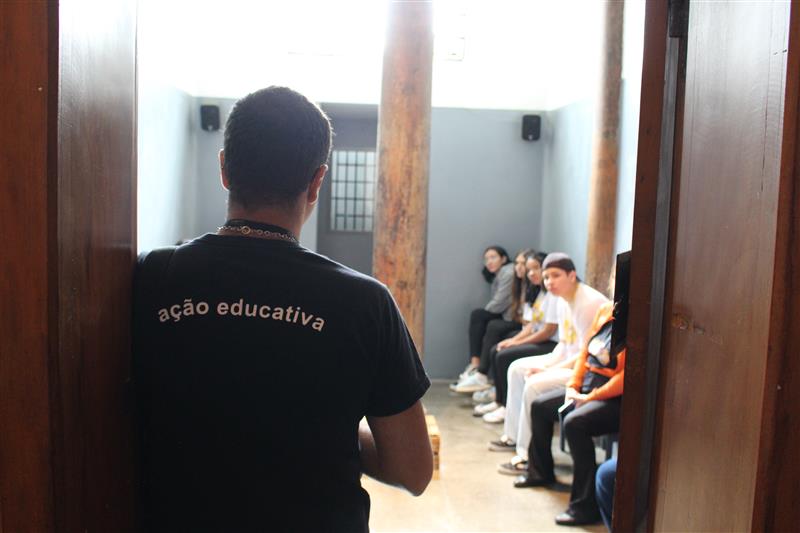 Um educador, visto de costas, está parado na entrada de uma cela iluminada por luz natural. Ele usa uma camiseta preta com a frase “ação educativa” nas costas. À sua frente, várias pessoas estão sentadas em um banco encostado na parede, ouvindo ou aguardando instruções.