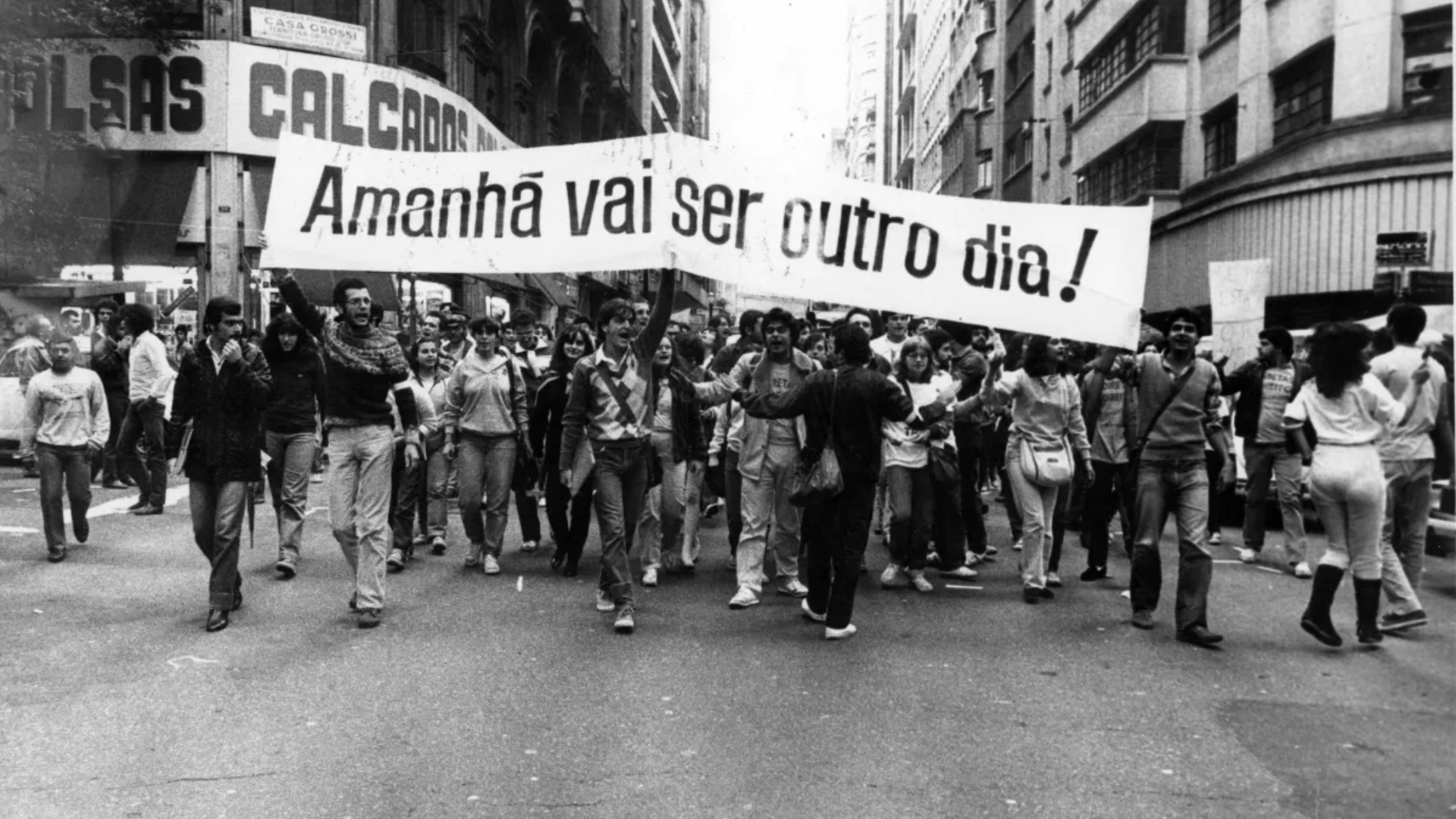 Fotografia em preto e branco de uma manifestação em uma rua do centro da cidade. Um grupo grande de pessoas caminha segurando uma faixa longa com a frase ‘Amanhã vai ser outro dia!’, ocupando toda a largura da via.