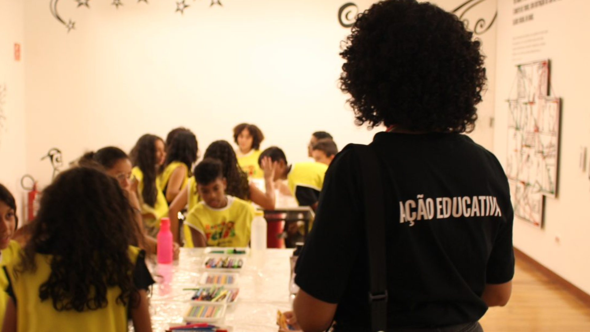 Educador acompanha uma atividade de desenho em uma sala com várias crianças reunidas ao redor de uma mesa com materiais artísticos.
