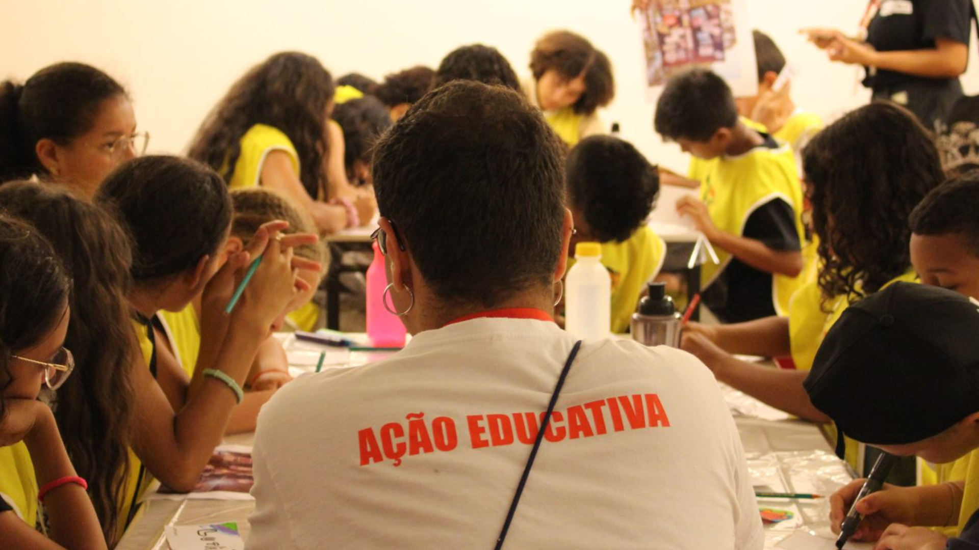 Pessoa com camiseta escrita ‘Ação Educativa’ acompanha um grupo de crianças reunidas ao redor de uma mesa participando de uma atividade de desenho