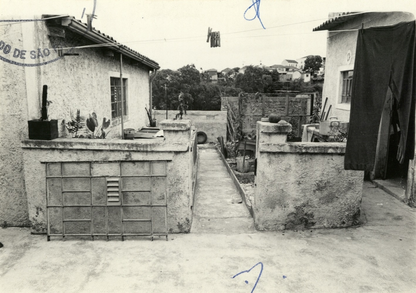Foto em preto e branco do quintal da Casa da Lapa. Mostra o espaço cimentado entre a casa principal ( a esquerda) e a dos fundos (a direita). Da esquerda para direita uma mureta de aproximadamente dois metros de largura, um corredor e outra mureta.